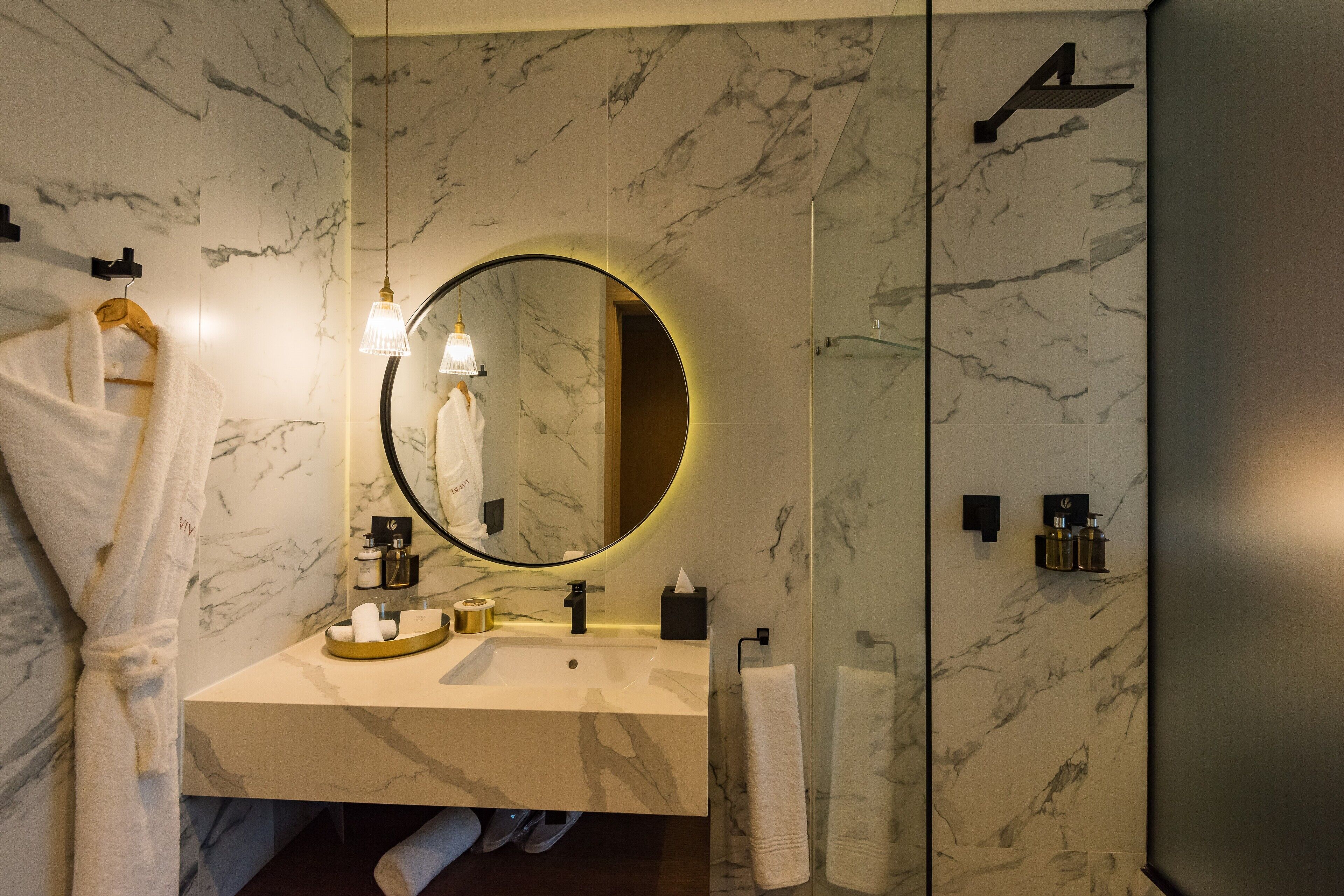 A modern bathroom with a marble backsplash, a round mirror, a sink with toiletries, and a towel rack.