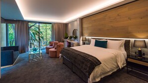 A modern hotel room with a large bed, a seating area with a sofa and chairs, a small table, and a view of greenery through the window.