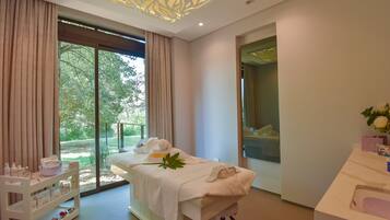 Relax in a serene spa room with a sliding glass door opening to a lush garden view, featuring a massage table, reception desk, and shelving unit.