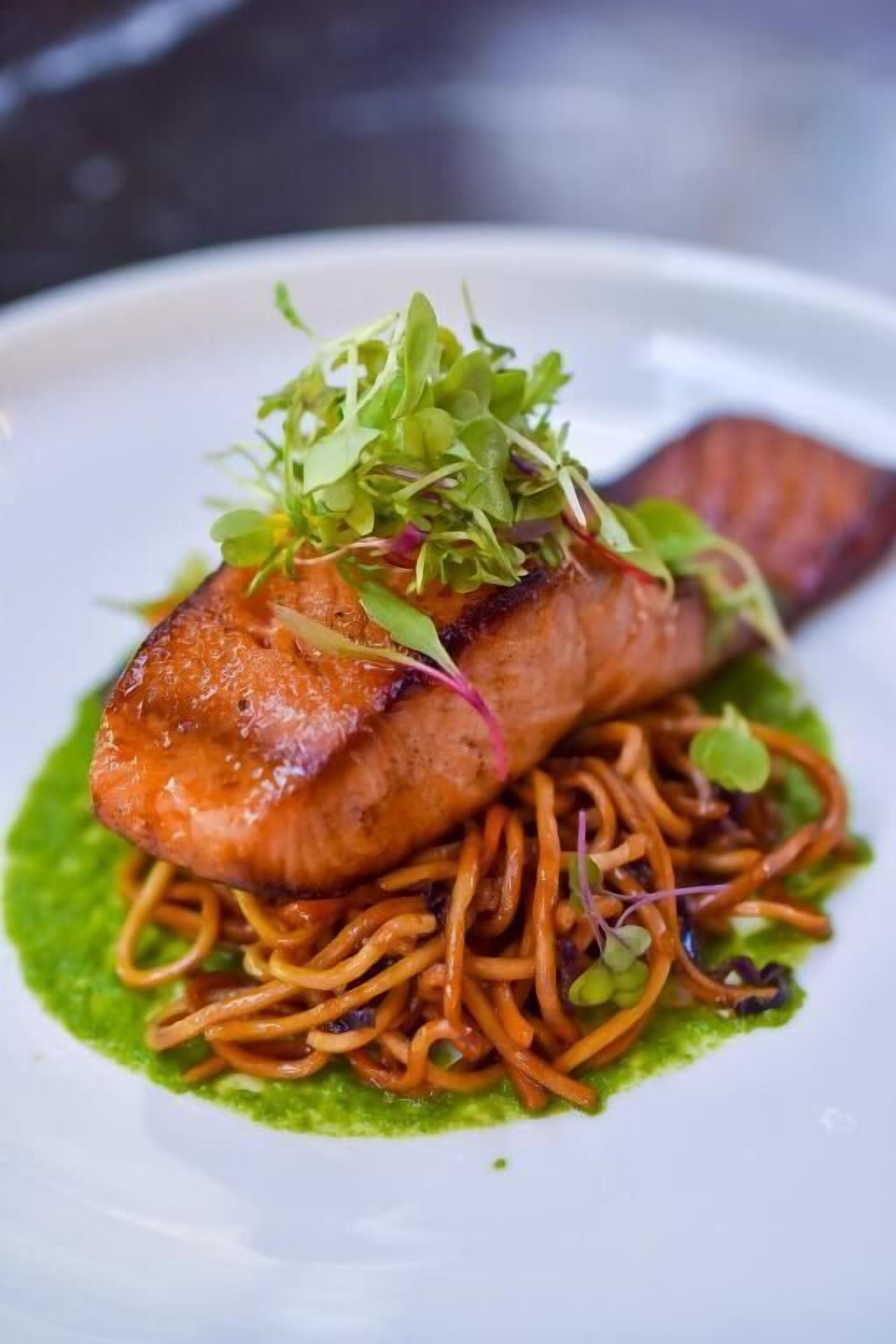 A plate with a piece of salmon, noodles, and a green sauce.