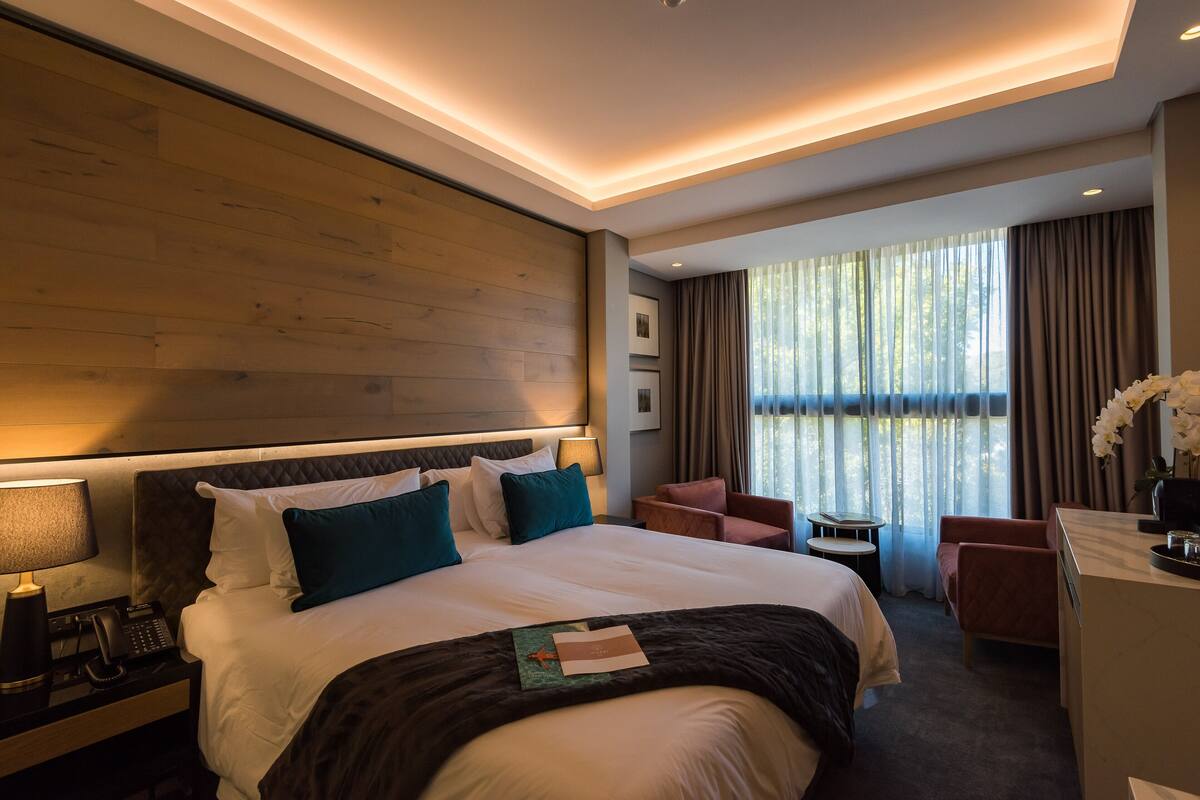 Ein modernes Hotelzimmer mit einem großen Bett, einem Schreibtisch, einem Stuhl und Blick ins Freie durch große Fenster.