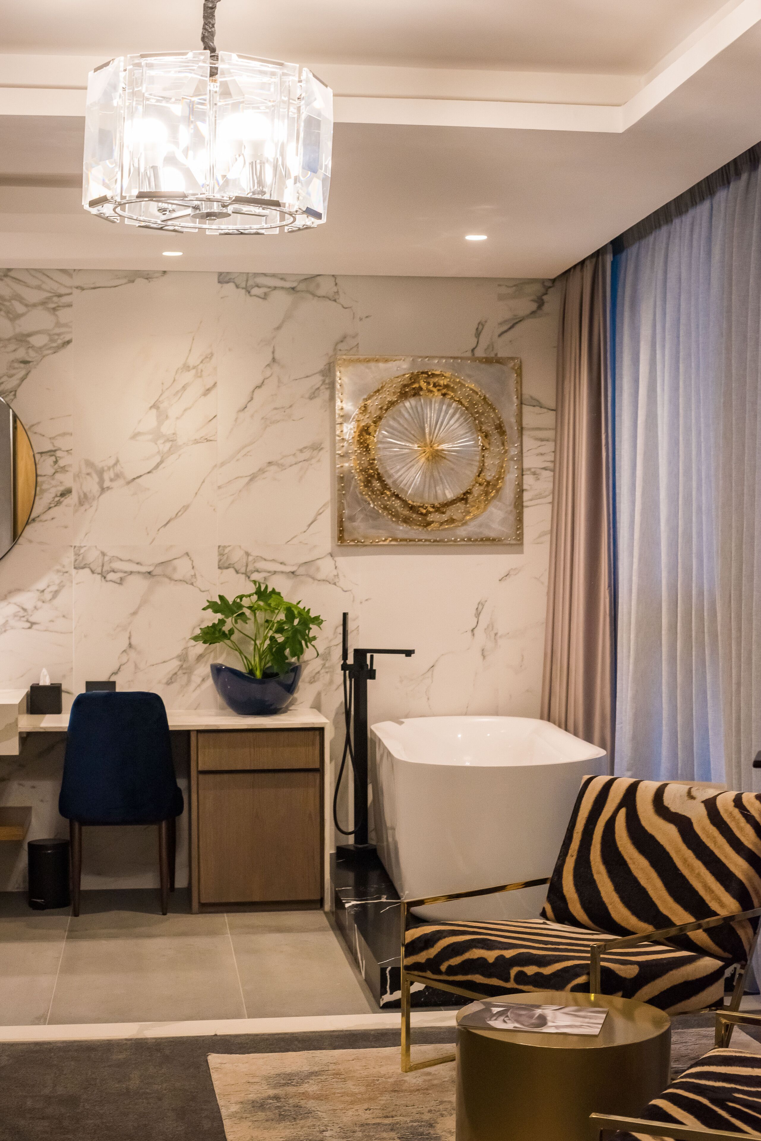 A modern bathroom with a marble wall, a large bathtub, a zebra-patterned chair, and a blue vase with green plants.