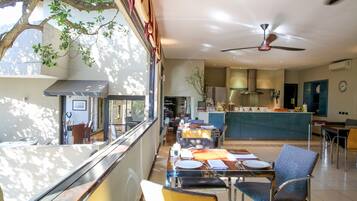 A dining area with tables and chairs, a kitchen in the background, and a ceiling fan.