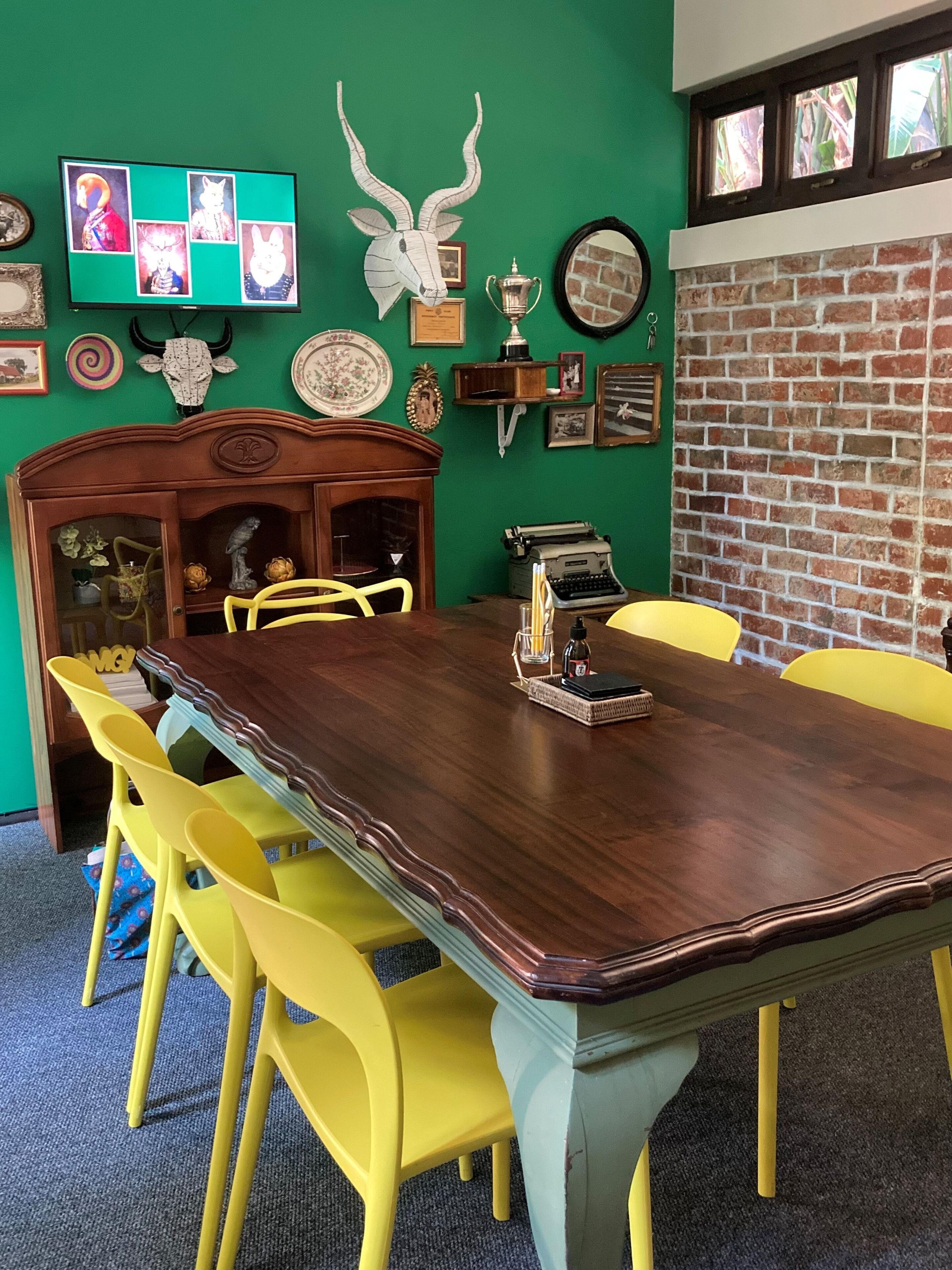 A dining area with a long wooden table, yellow chairs, and a green wall adorned with various decorations.