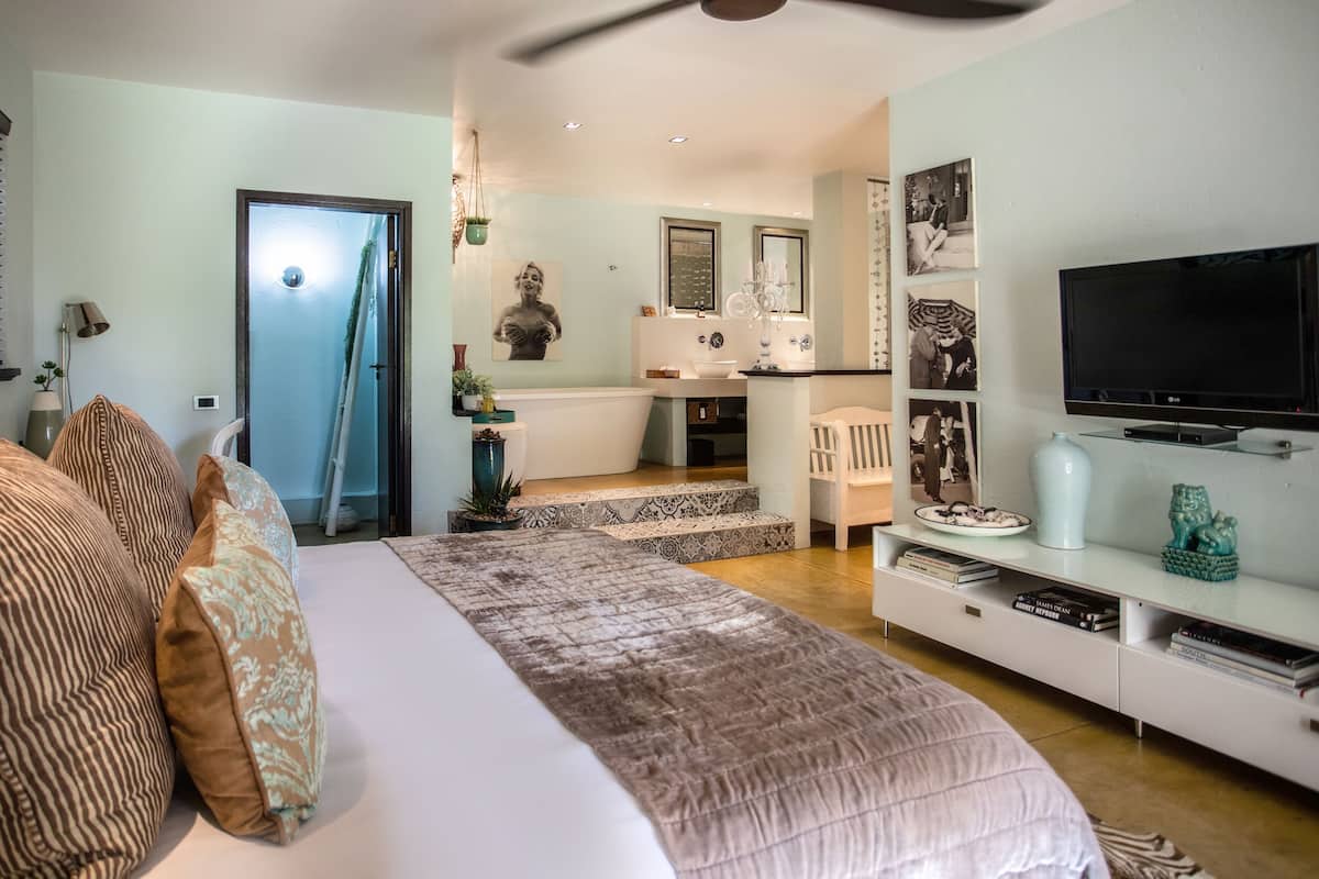 A bedroom with a bed, pillows, a TV, a bookshelf, and a bathroom visible in the background.
