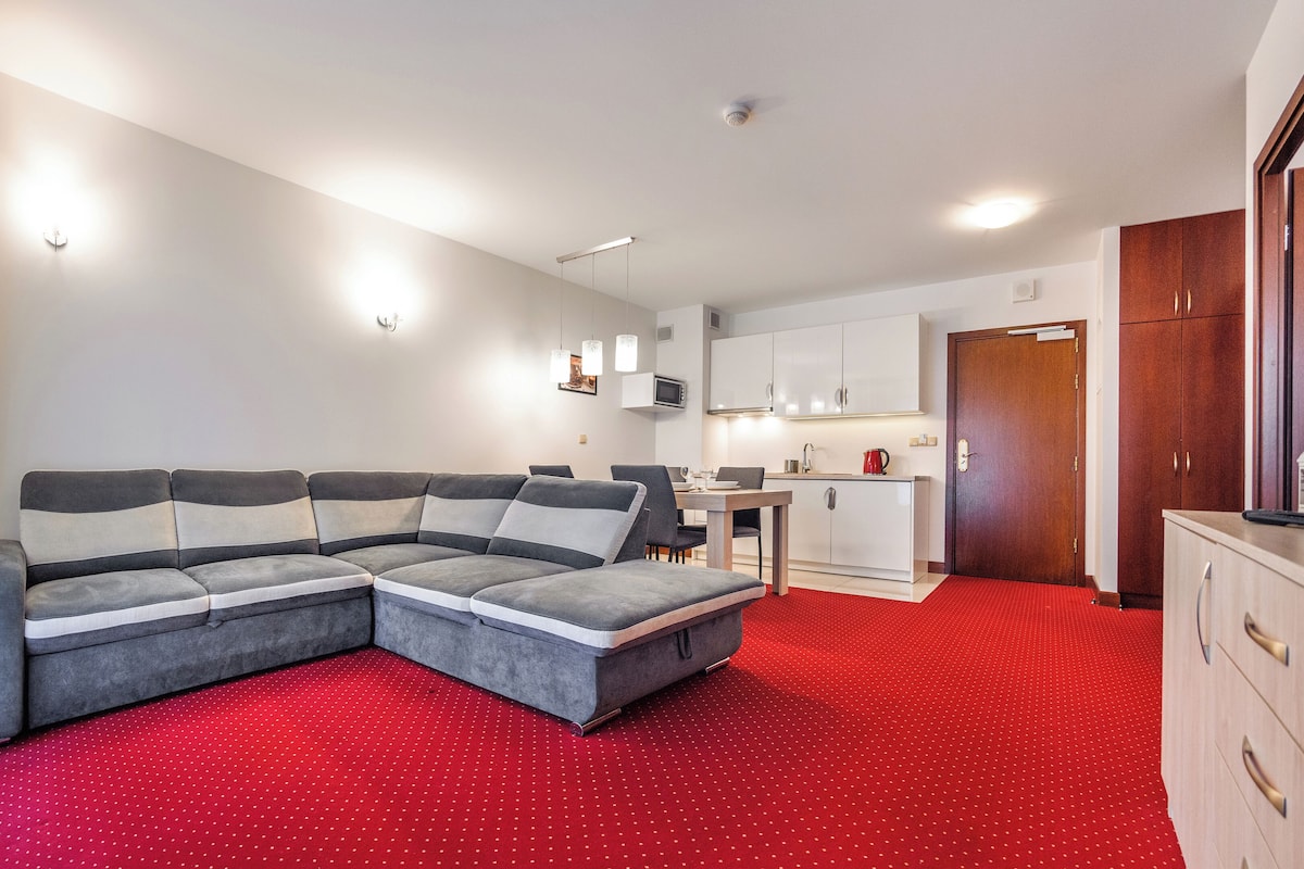 Una sala moderna con un sofá modular, una zona de comedor y una cocina al fondo, además de una alfombra roja.