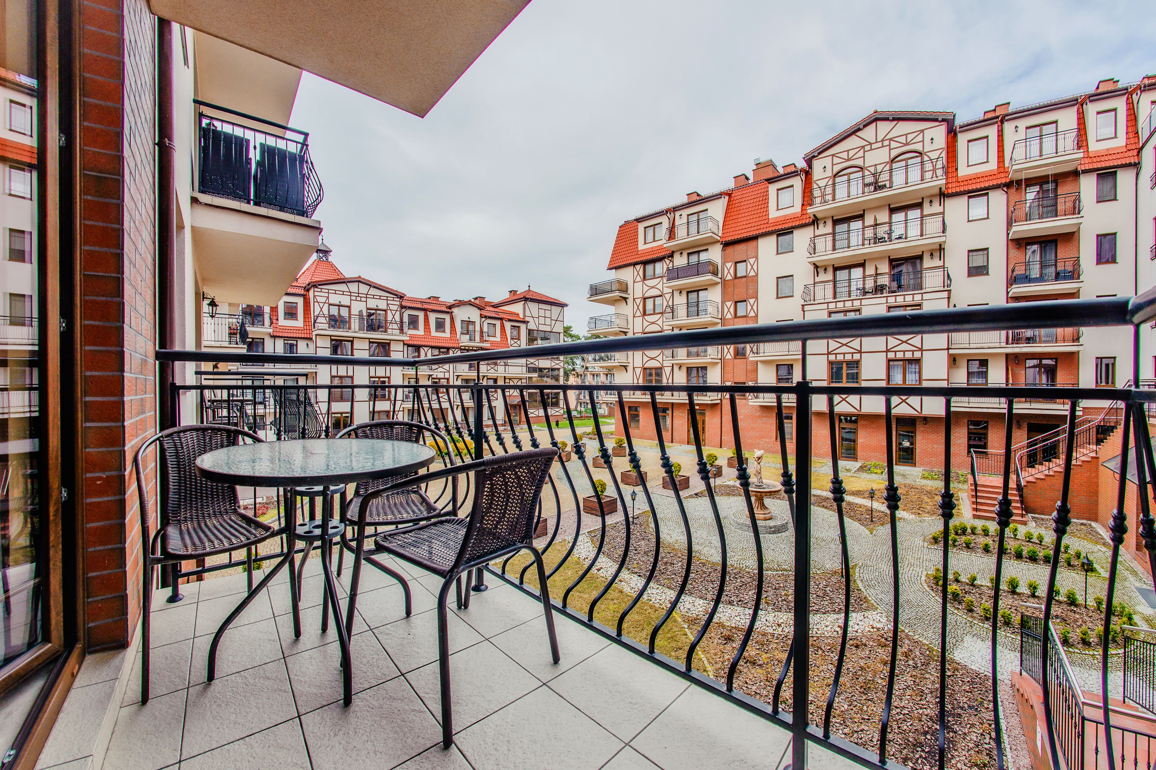 Uma varanda com mesa e cadeiras, com vista para um pátio com edifícios e uma escadaria.