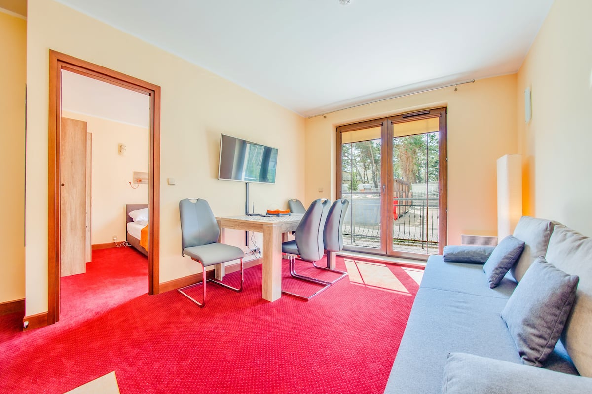 Habitación de hotel con alfombra roja, un sofá gris, un televisor montado en la pared y un balcón con puerta de cristal.