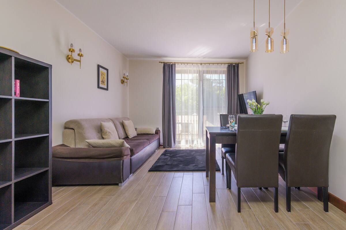 Una sala de estar con sofá, una mesa de comedor con sillas, una estantería y una ventana con cortinas.