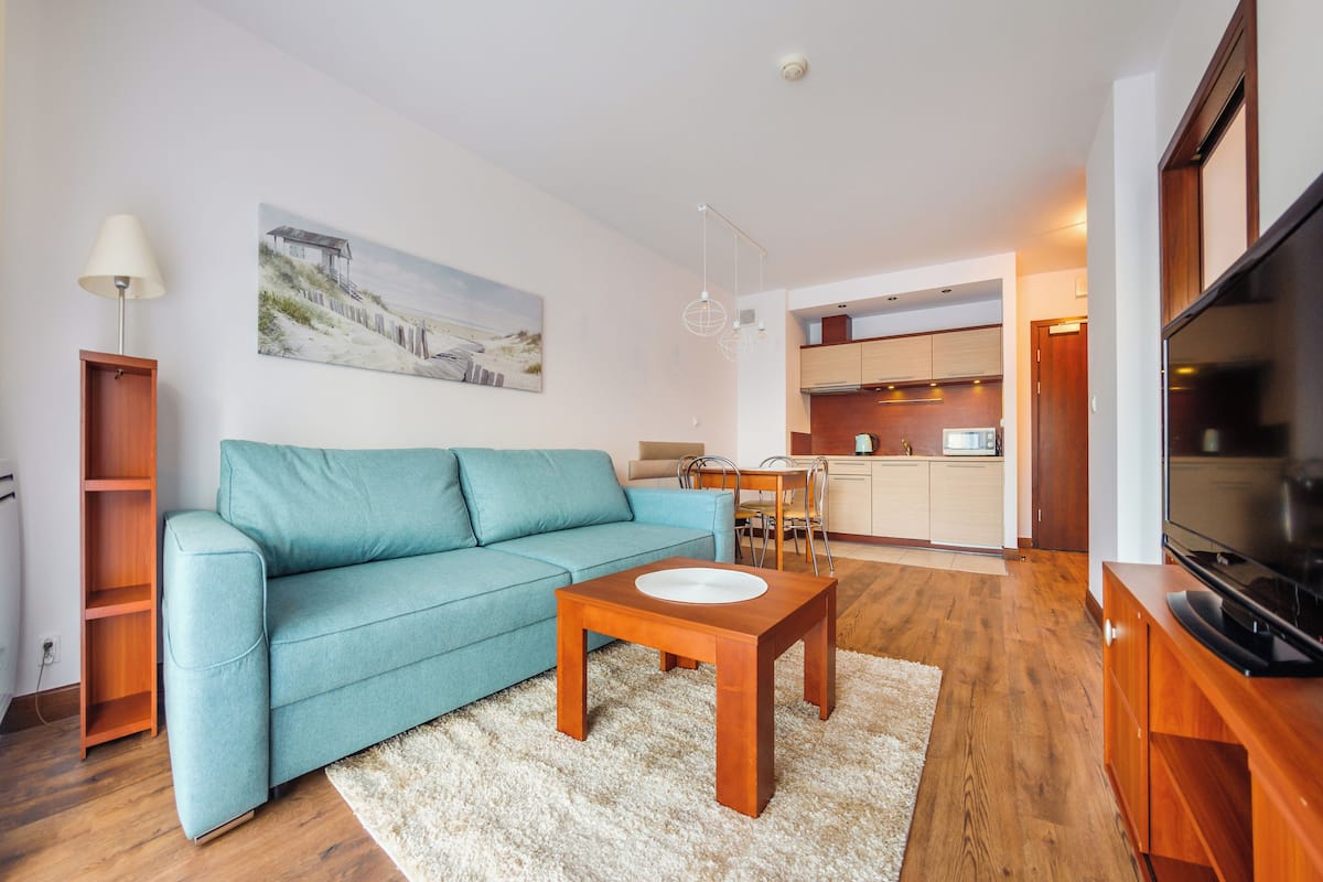 Una sala moderna con un sofá color turquesa, una mesa de centro de madera, un televisor y una cocina al fondo.