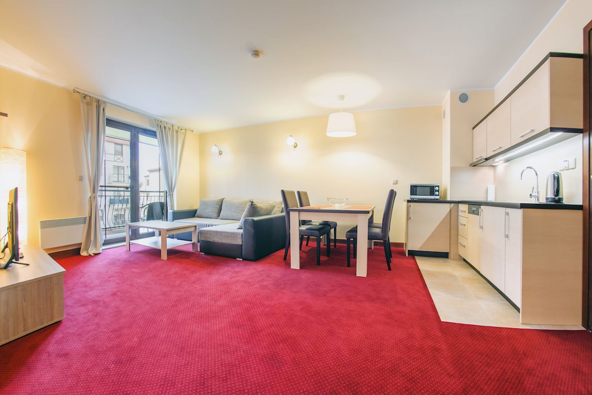 Un espacio habitable compacto con alfombra roja, un sofá, una mesa de comedor y una kitchenette.