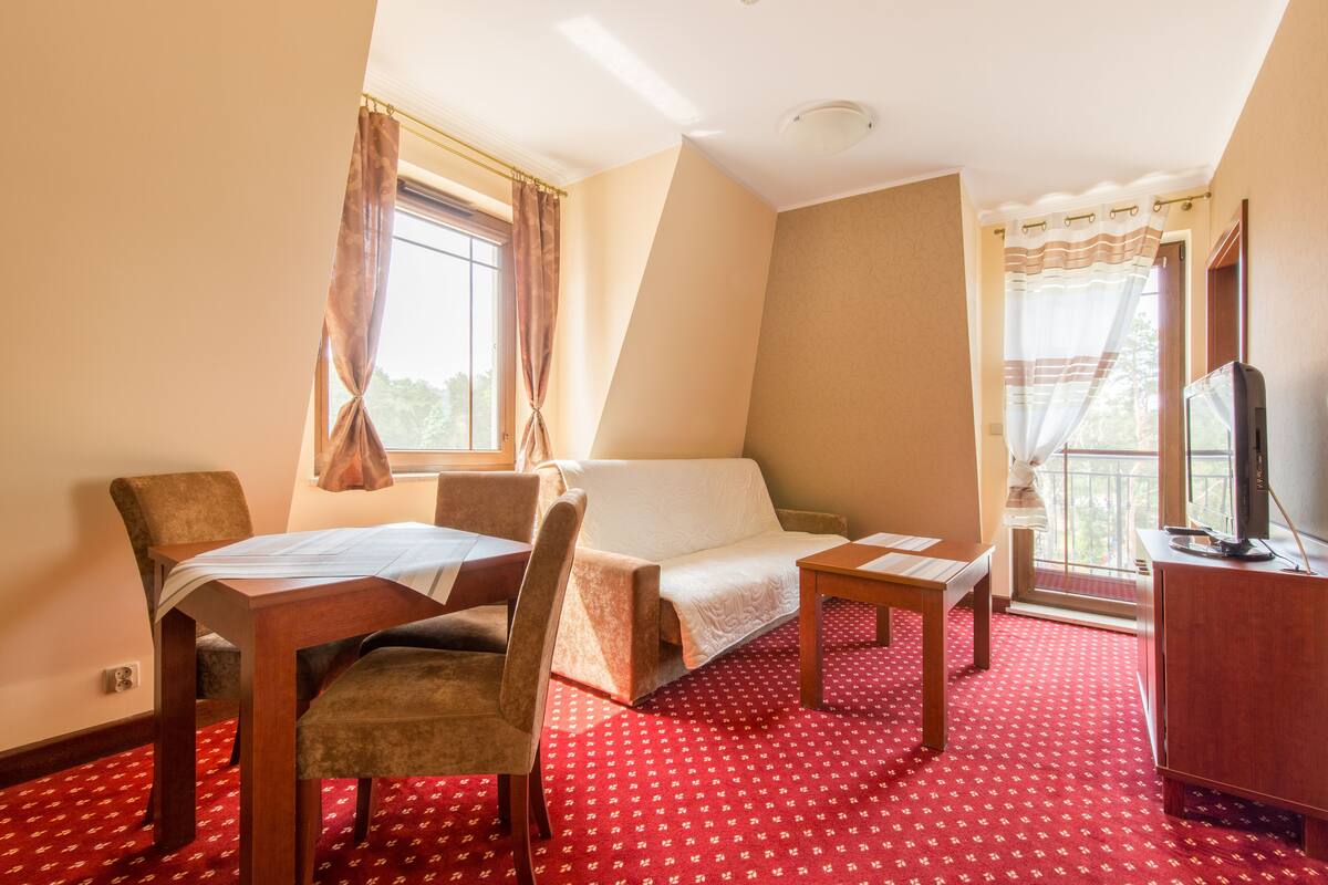 Habitación de hotel con cama, escritorio, silla, mesita pequeña, televisor y ventana con cortinas.