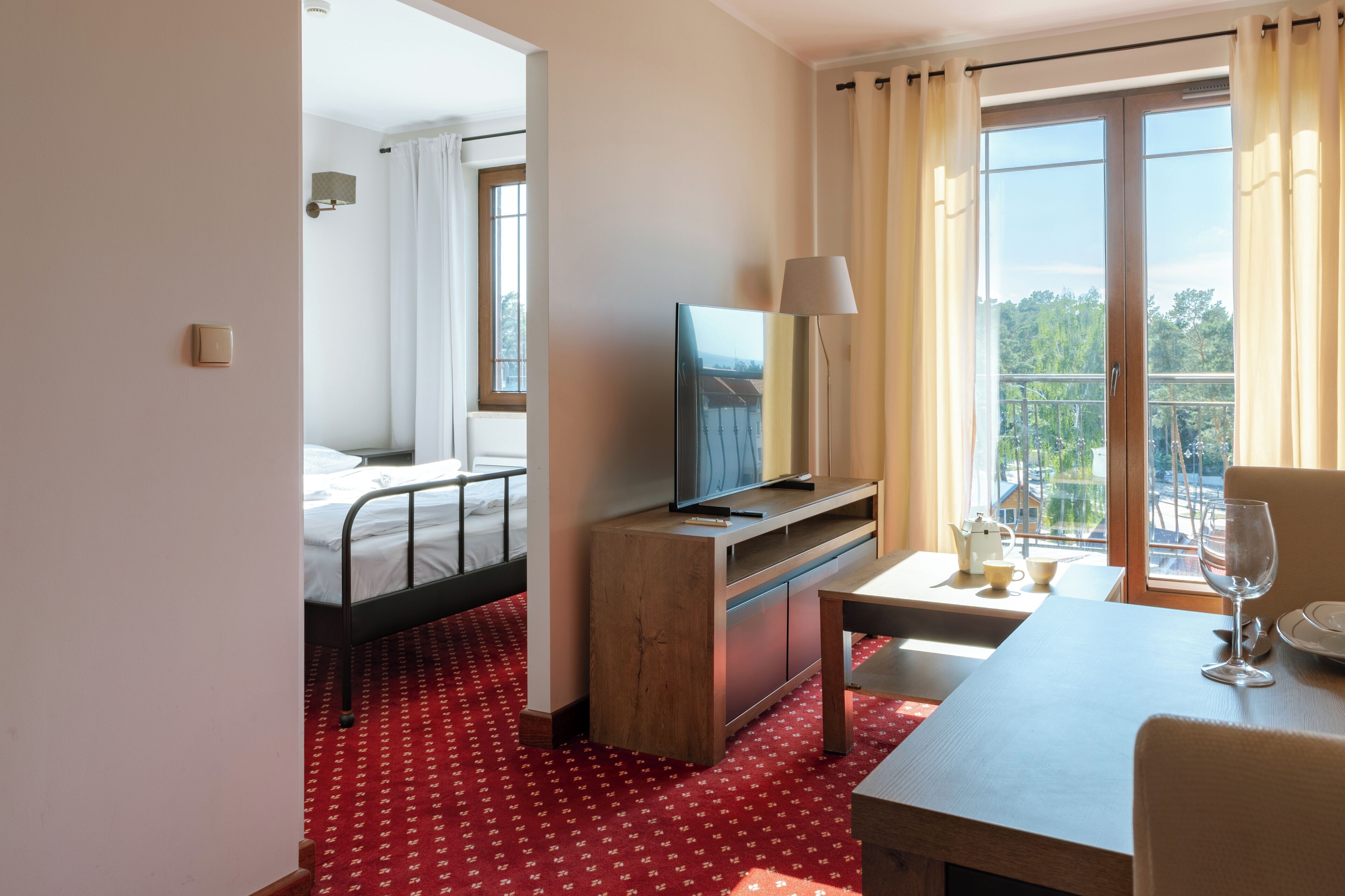Une chambre d’hôtel avec un lit, un téléviseur posé sur un meuble en bois, une table à manger avec un verre, et un balcon avec des rideaux.