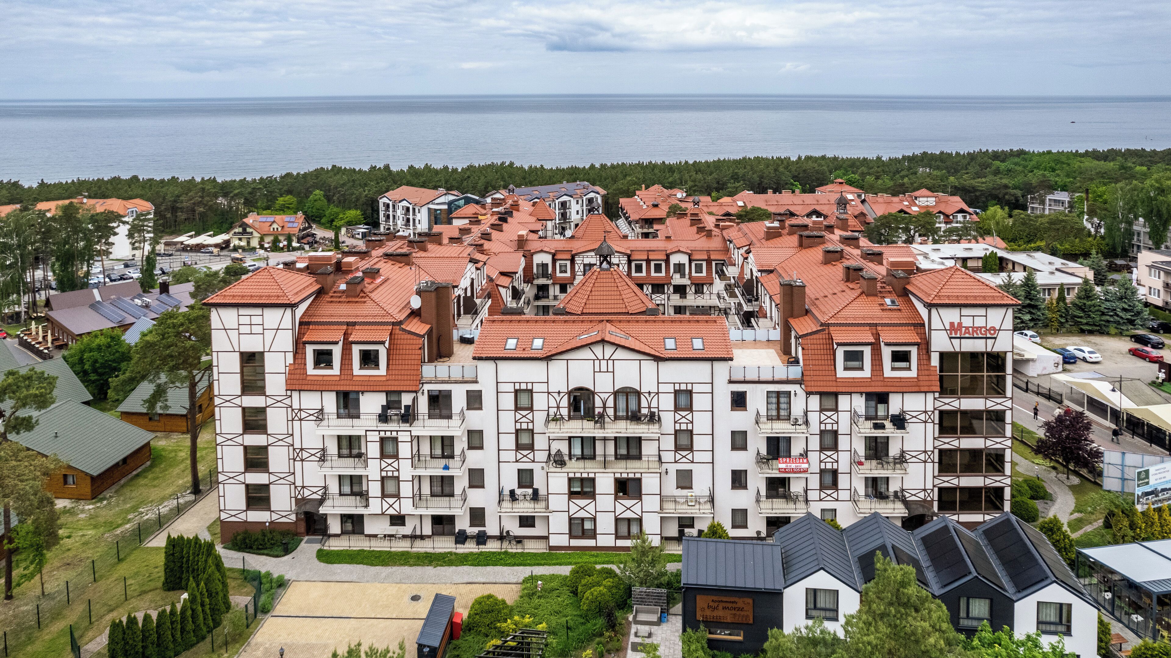 A large, multi-story building with a red-tiled roof, surrounded by greenery and a view of the sea.
