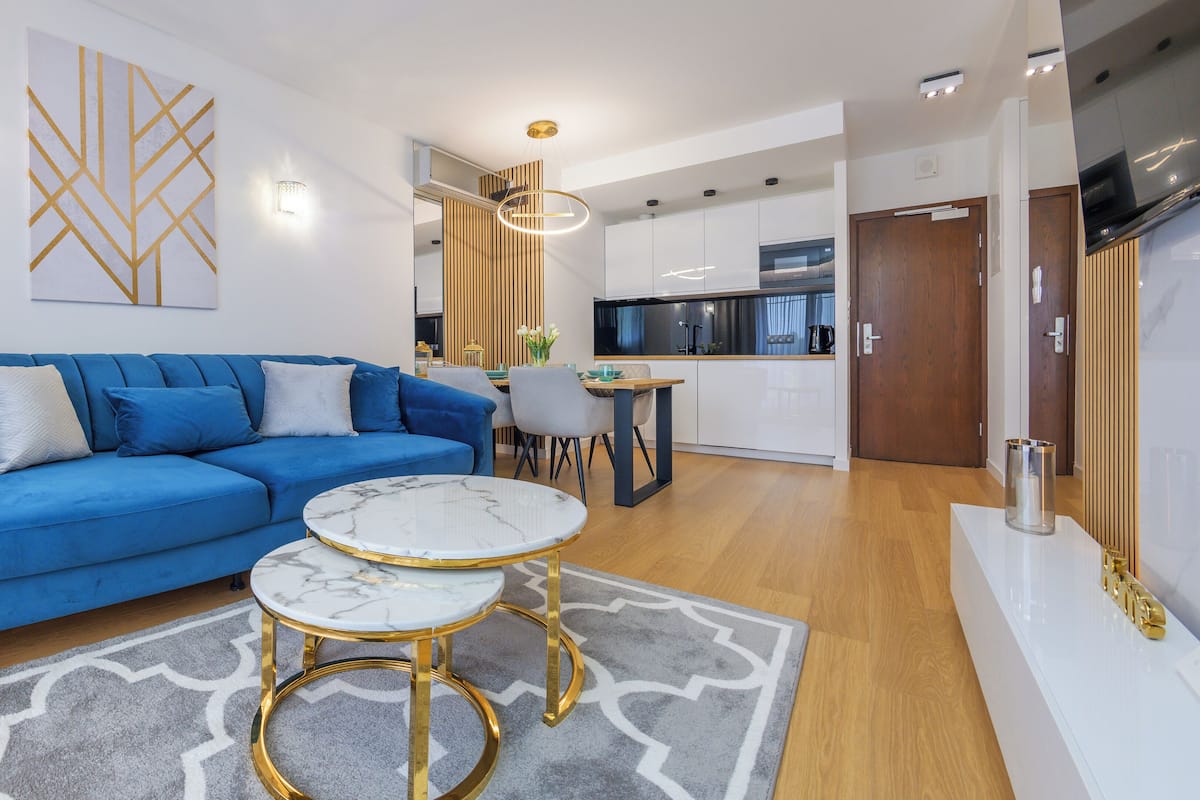 Una sala de estar moderna con un sofá azul, una mesa de centro de mármol y una cocina al fondo.