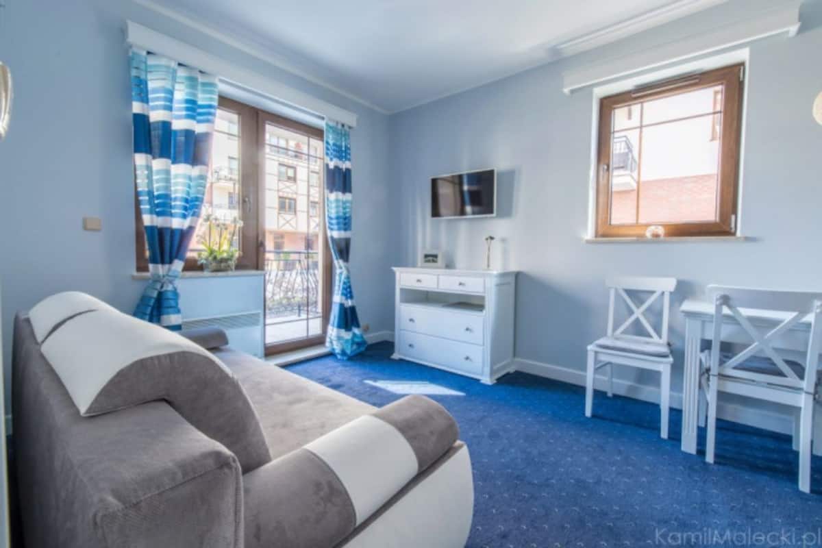 Una habitación con un sofá gris, un tocador blanco, un televisor en la pared y dos sillas blancas.