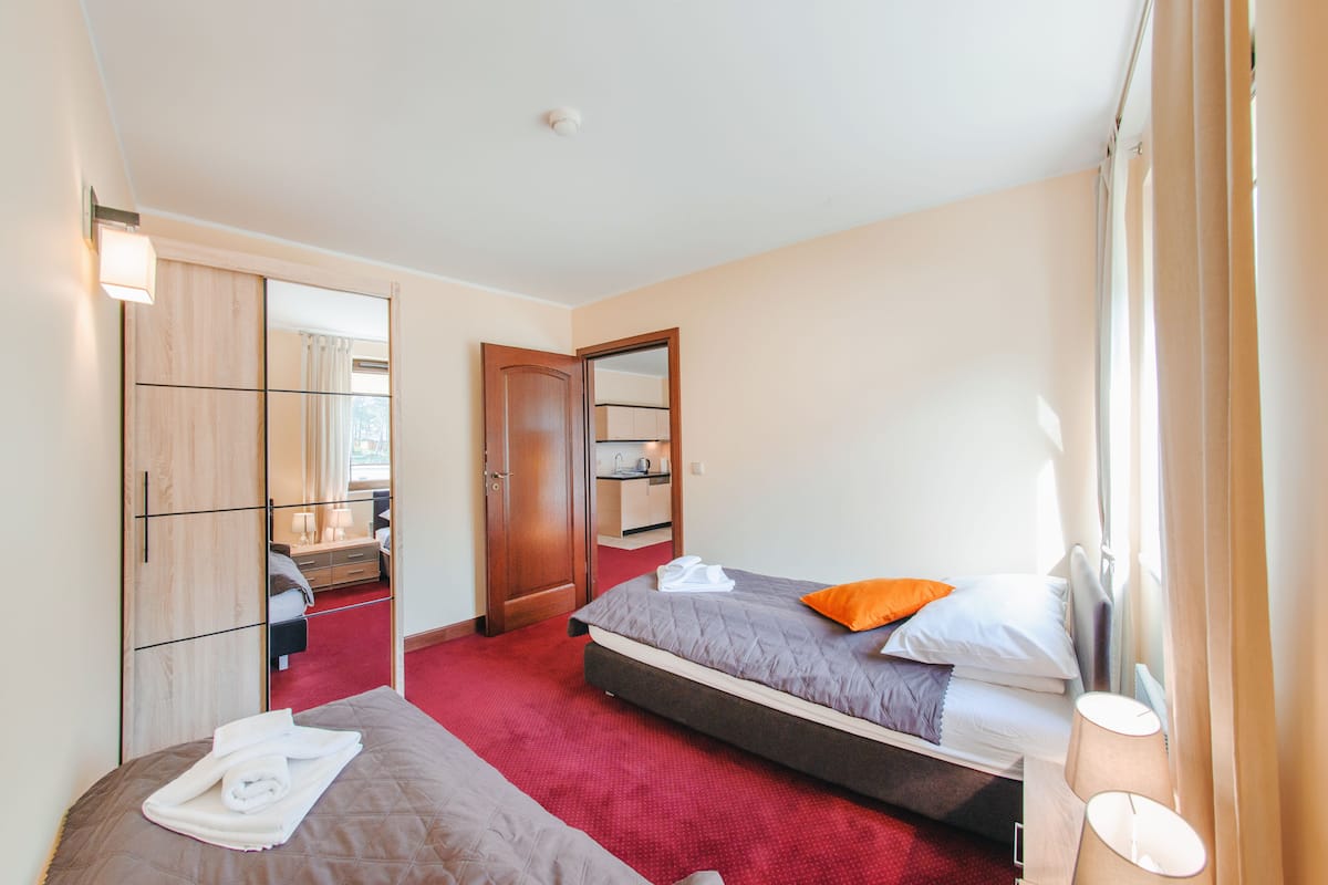 Habitación de hotel con dos camas, alfombra roja, armario de madera y baño a la vista a través de una puerta abierta.