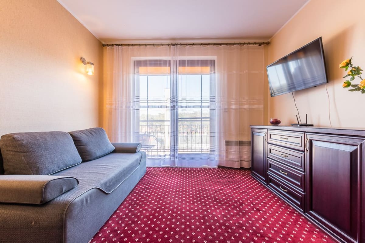 Una habitación de hotel con un sofá gris, una alfombra roja estampada, un mueble de madera oscura, un televisor de pantalla plana y una ventana con cortinas translúcidas.