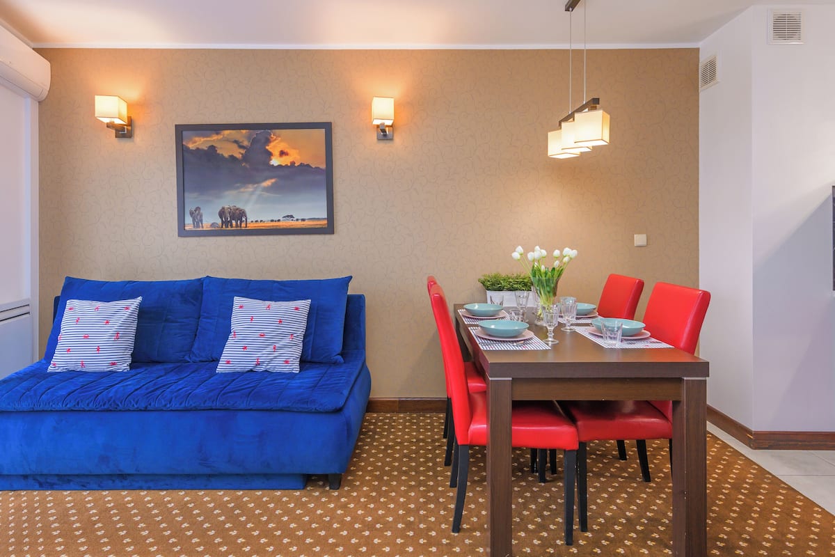 Una habitación moderna con sofá azul, sillas rojas, una mesa de comedor y una zona de cocina.