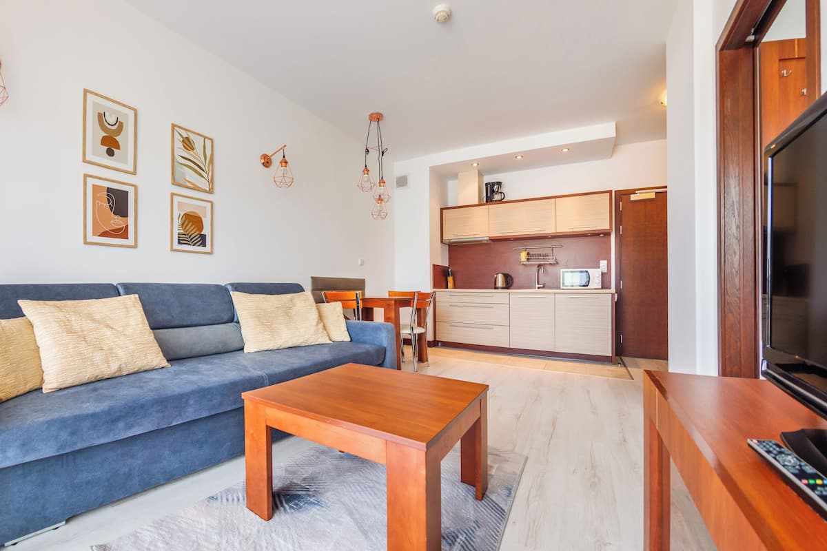 Una sala de estar con un sofá azul, una mesa de centro de madera y un mueble para televisor. Hay una zona de cocina con armarios y un microondas.