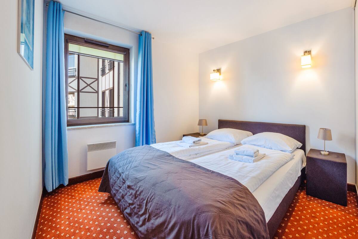 Una cama bien hecha con sábanas blancas, una ventana con cortinas azules y dos lámparas de pared.