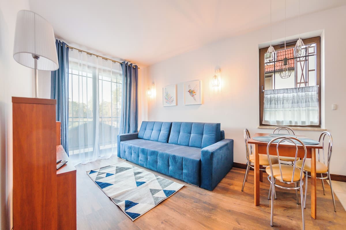 Una sala de estar con un sofá azul, una mesa de comedor de madera con sillas, un tapete estampado y un ventanal con cortinas.