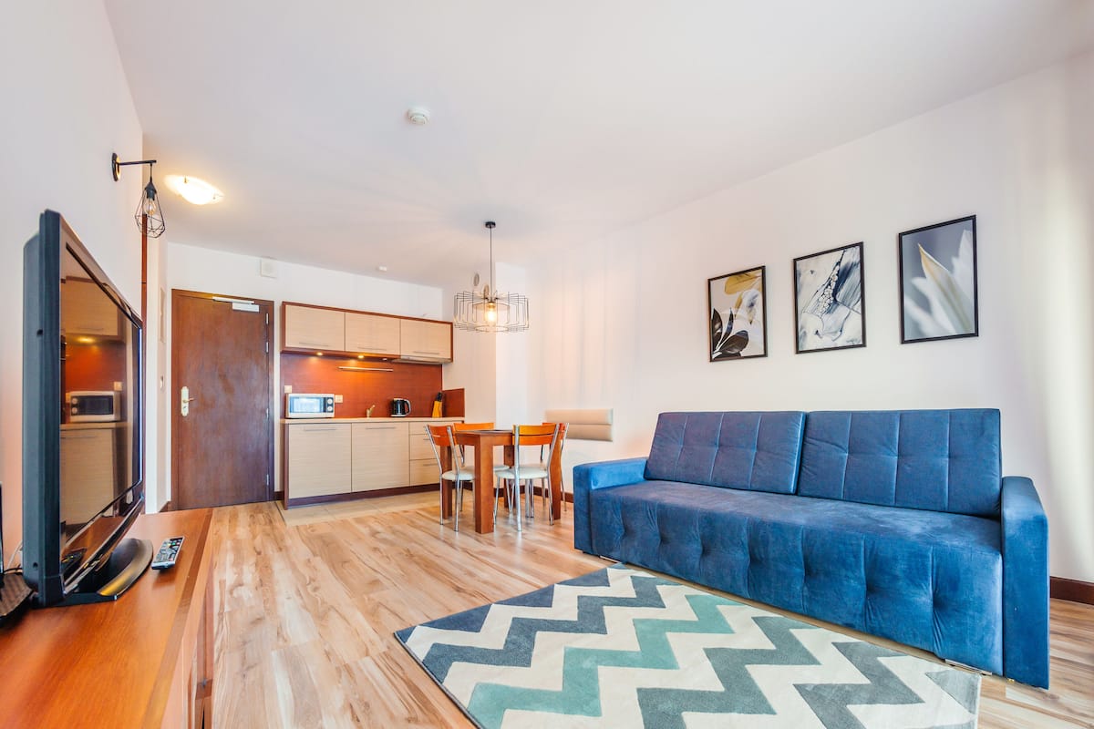 Una sala de estar moderna con un sofá azul, piso de madera, una zona de comedor con una mesa y sillas, y una cocina al fondo.
