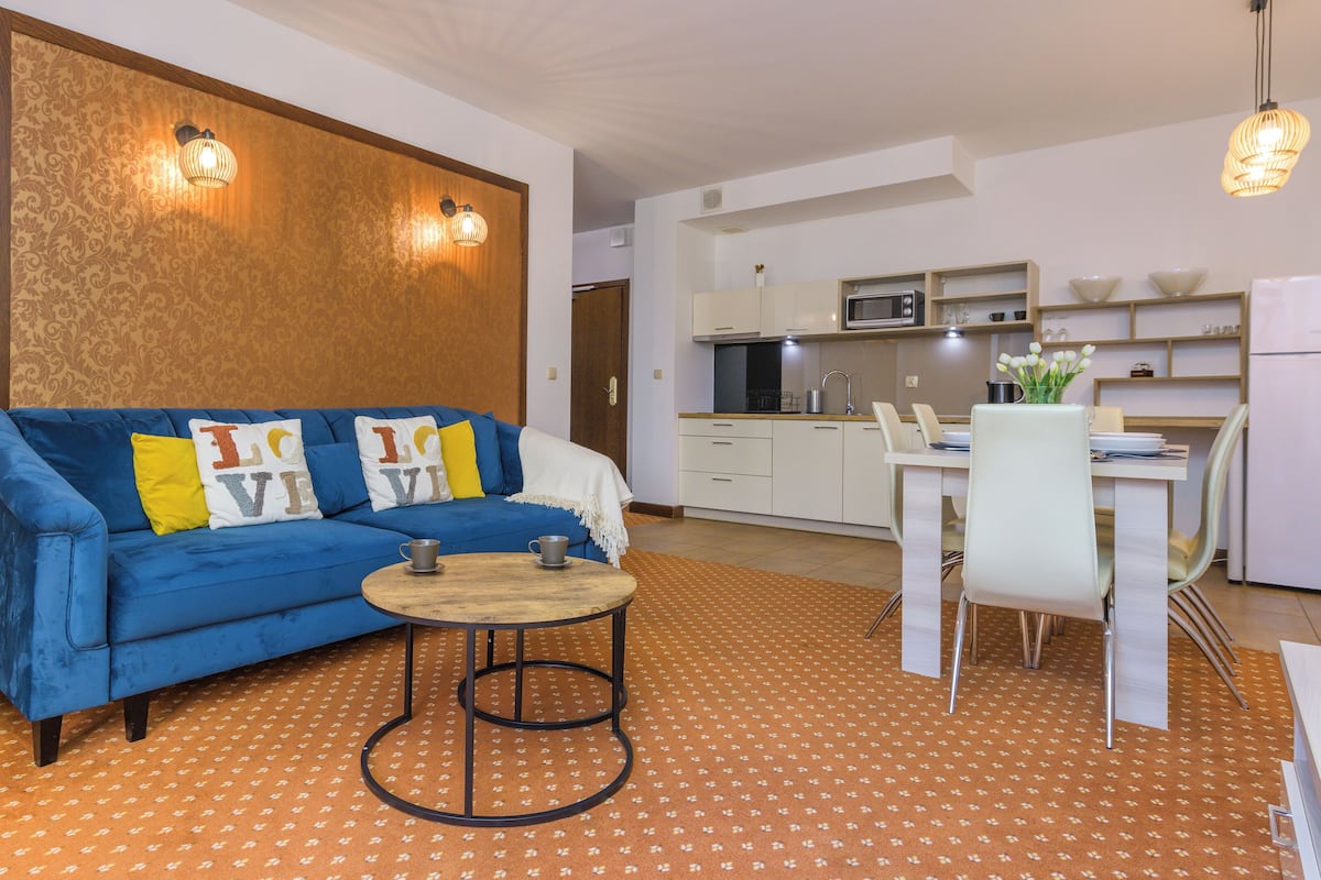 Una sala moderna con un sofá azul, una mesita de madera y una cocina con armarios blancos y una mesa de comedor.