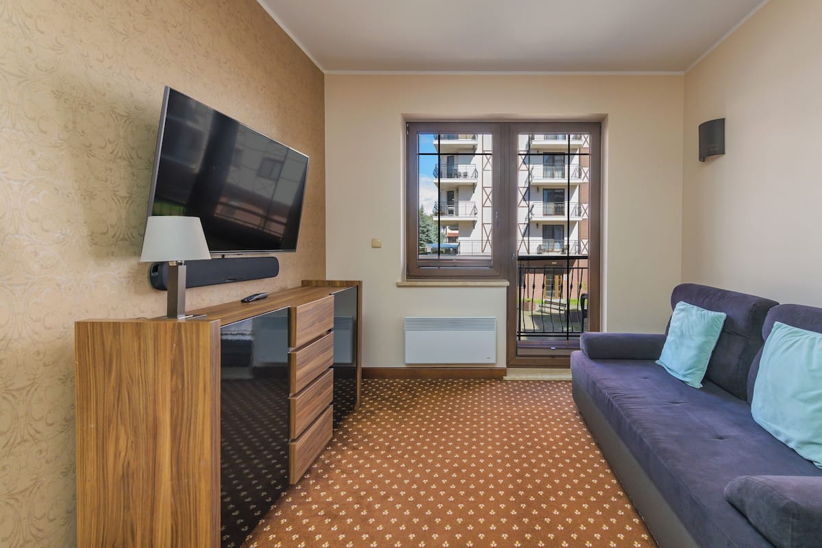 Un salón moderno con un televisor de pantalla plana, un mueble de madera, un sofá con cojines, una alfombra estampada y una ventana con vistas a un balcón.