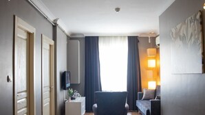 A modern living room with a wooden floor, a dark armchair, a white side table, a sofa, and a large window with curtains. - The Hera Suit Hotels (Istanbul)