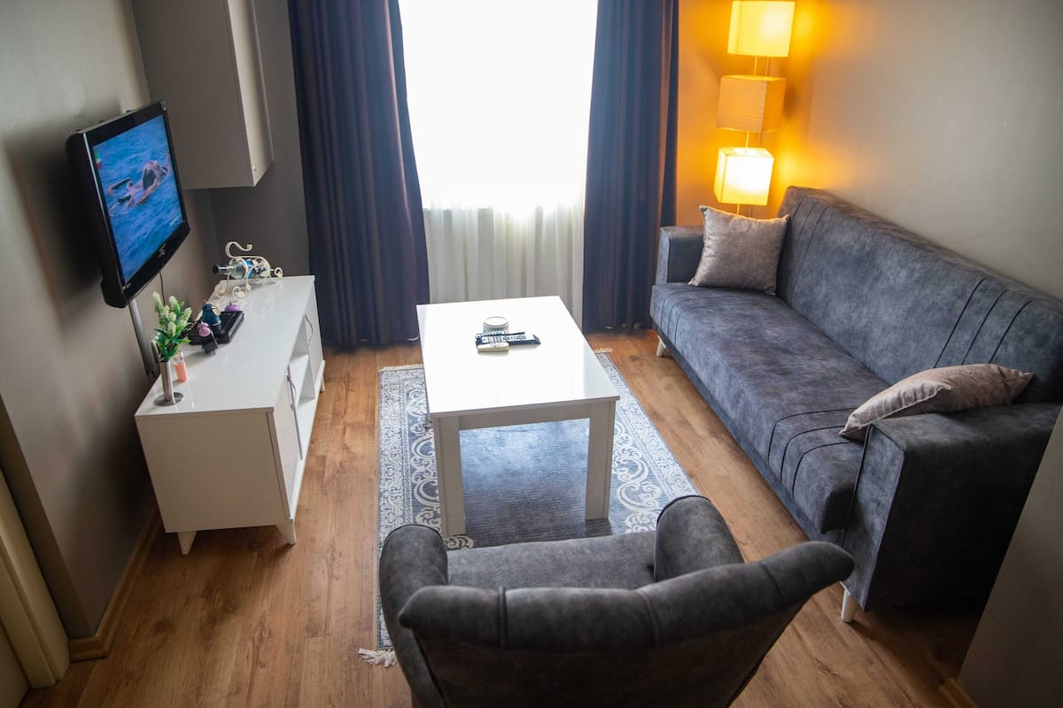 Un salón con un sofá gris, una mesa de centro blanca, un televisor en la pared y una lámpara auxiliar.