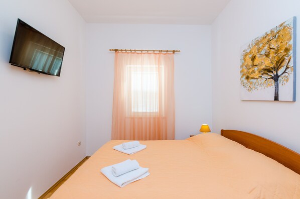Une chambre d’hôtel avec un lit, un téléviseur fixé au mur, une fenêtre avec des rideaux légers et un tableau représentant un arbre.