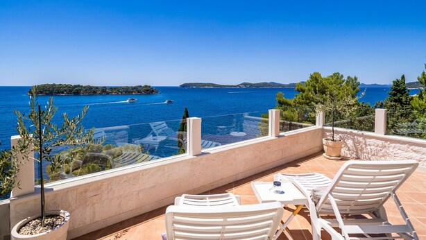 A terrace with white chairs and a table, overlooking a clear blue sea with boats and a distant island.