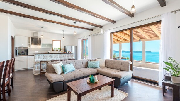 A spacious living area with a large sectional sofa, a wooden coffee table, and a dining set. A kitchen with modern appliances is visible in the background.
