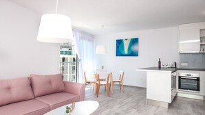 A modern living room with a pink sofa, a white coffee table, and a dining area with wooden chairs. A kitchen with white cabinets and an oven is visible in the background. - Dva Galeba - Comfort One Bedroom Apartment with Sea View (205) (Bol)