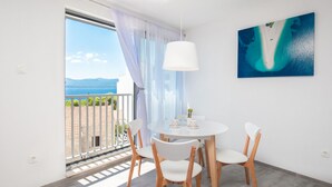 A room with a round table and chairs, a ceiling light, a painting of a beach, and a view of the sea through a window. - Dva Galeba - Comfort One Bedroom Apartment with Sea View (205) (Bol)