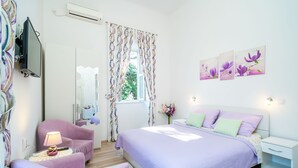 Ein Schlafzimmer mit Bett, Sessel, Nachttisch, Lampe, Fernseher und einem Fenster mit Vorhängen.