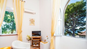 Ein kleiner, heller Raum mit Fernseher, einem weißen Sofa, einem kleinen Tisch und einem Fenster mit Blick auf Bäume.