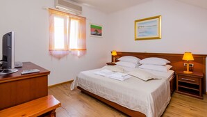 Uma cama bem arrumada com almofadas e um cobertor, uma mesa de cabeceira com abajur, uma TV sobre uma escrivaninha e uma janela com cortinas.