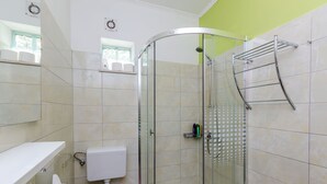 Casa de banho com chuveiro em cabine de vidro, sanita, lavatório com suporte para toalhas e prateleira com artigos de higiene pessoal.