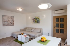 A modern living room with a sofa, a dining table with a fruit bowl, and two wall-mounted artworks.