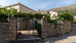 Une propriété aux murs de pierre, dotée d’une entrée fermée par un portail, un bâtiment en pierre avec un toit en tuiles, et une colline en arrière-plan.