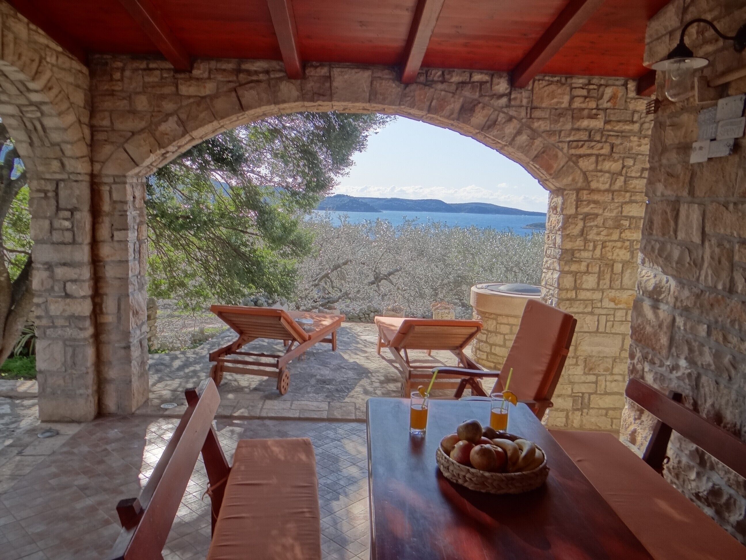 Un arco in pietra conduce a una terrazza con mobili in legno e un tavolo con frutta.