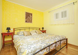 A bedroom with a bed, two bedside tables, a nightstand, a window with lace curtains, and a wall with a patterned wallpaper.