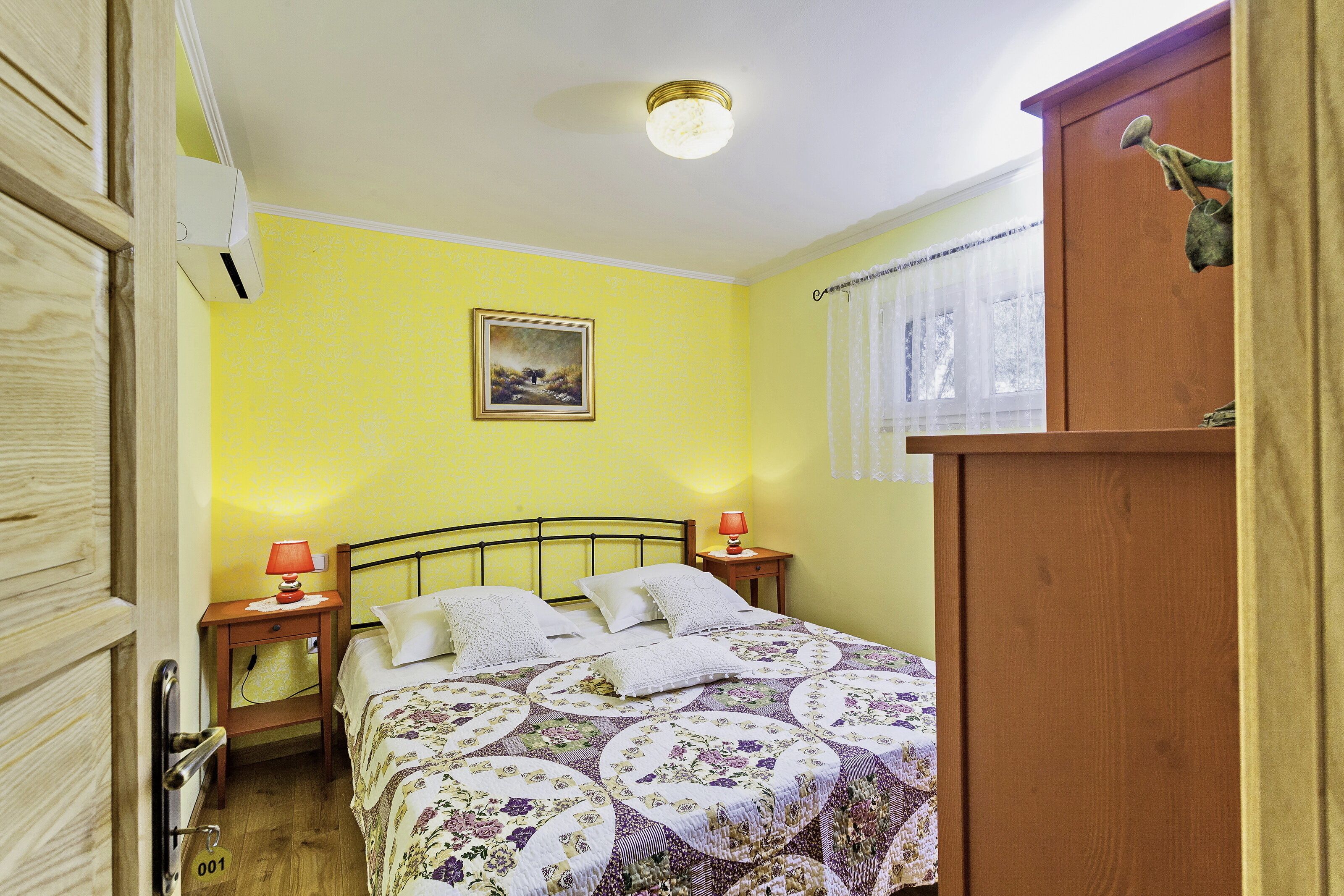 Une chambre comprenant un lit, deux tables de chevet, un climatiseur fixé au mur, une fenêtre avec des rideaux et une armoire en bois.