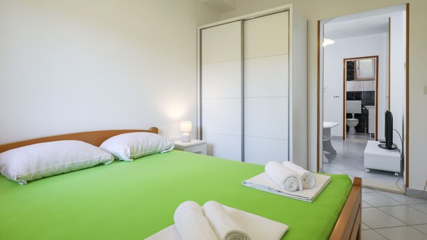 A hotel room with a green bed, white pillows, and a wooden headboard. There are towels on the bed and a nightstand with a lamp. A bathroom is visible through an open door. - Apartment Brane - Studio Apartment (Mokosica)
