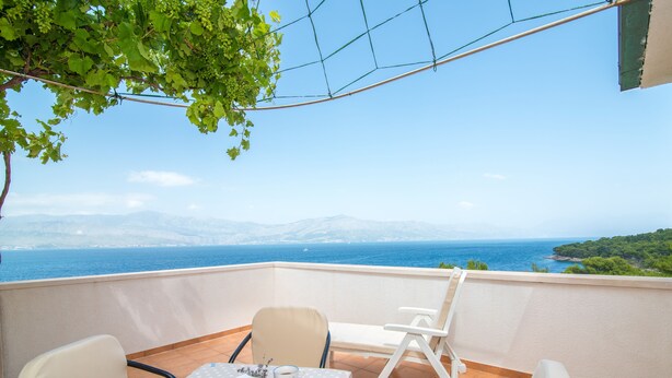 A balcony with a table and chairs, overlooking a coastal view with mountains in the distance. - Apartments Ana & Bogdan (Postira)
