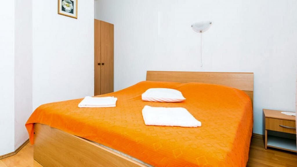 A neatly made bed with an orange bedspread and two white pillows, a wooden nightstand with a lamp, and a wooden wardrobe.