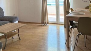 Ein Zimmer mit Holzboden, einer Couch, einem Esstisch mit Stühlen und einem großen Fenster mit Vorhängen.