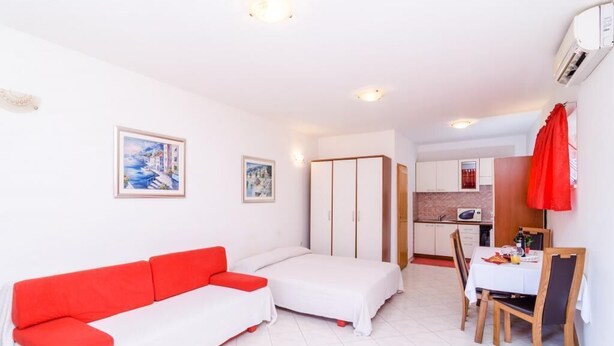 A compact room with a white sofa, a bed, a dining table with chairs, and a kitchen area.